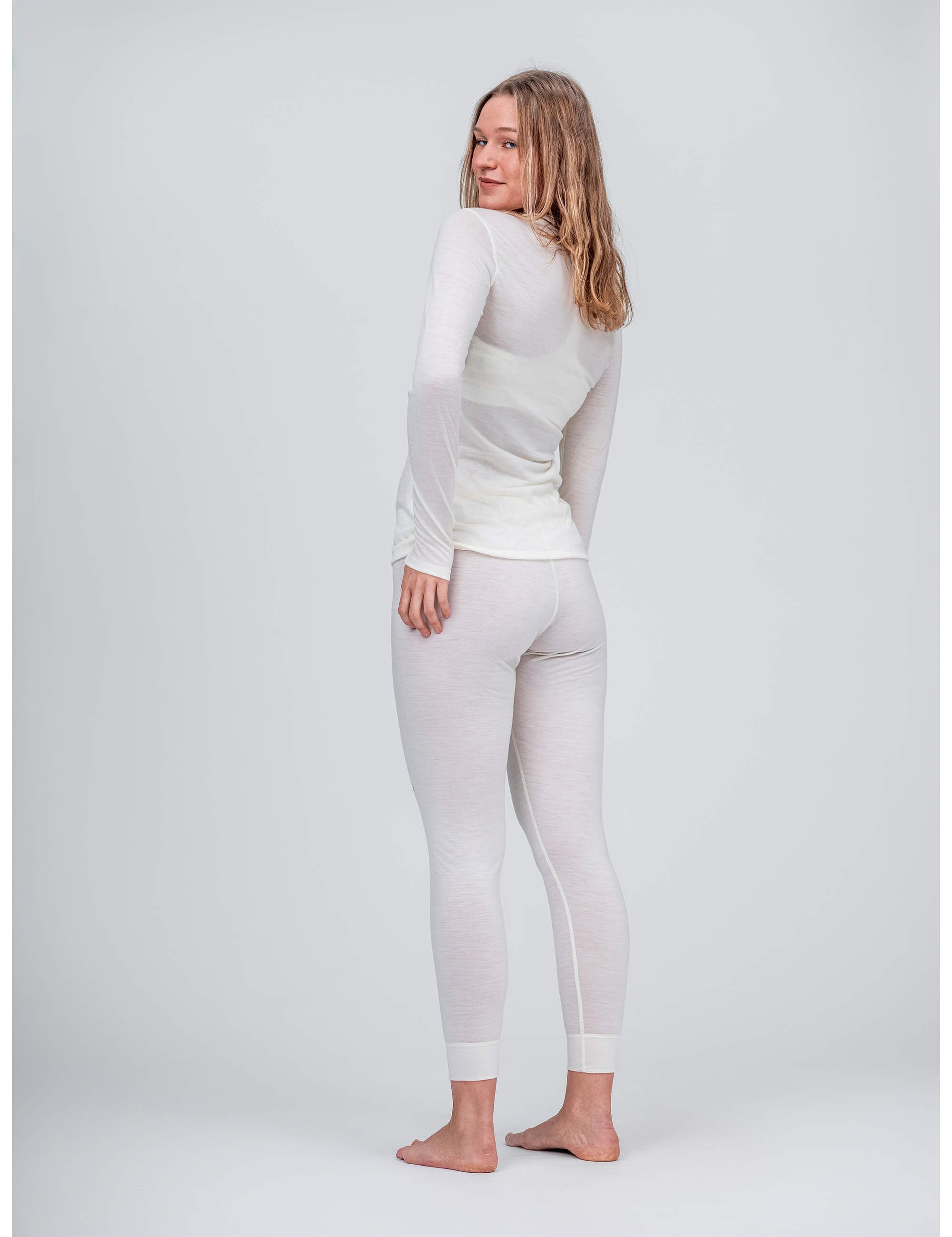 A woman stands barefoot on a light grey background, smiling over her shoulder and wearing the Crew Neck – Classic Merino 160 Women as a fitted, long-sleeve top with matching white leggings—perfect for everyday wear.