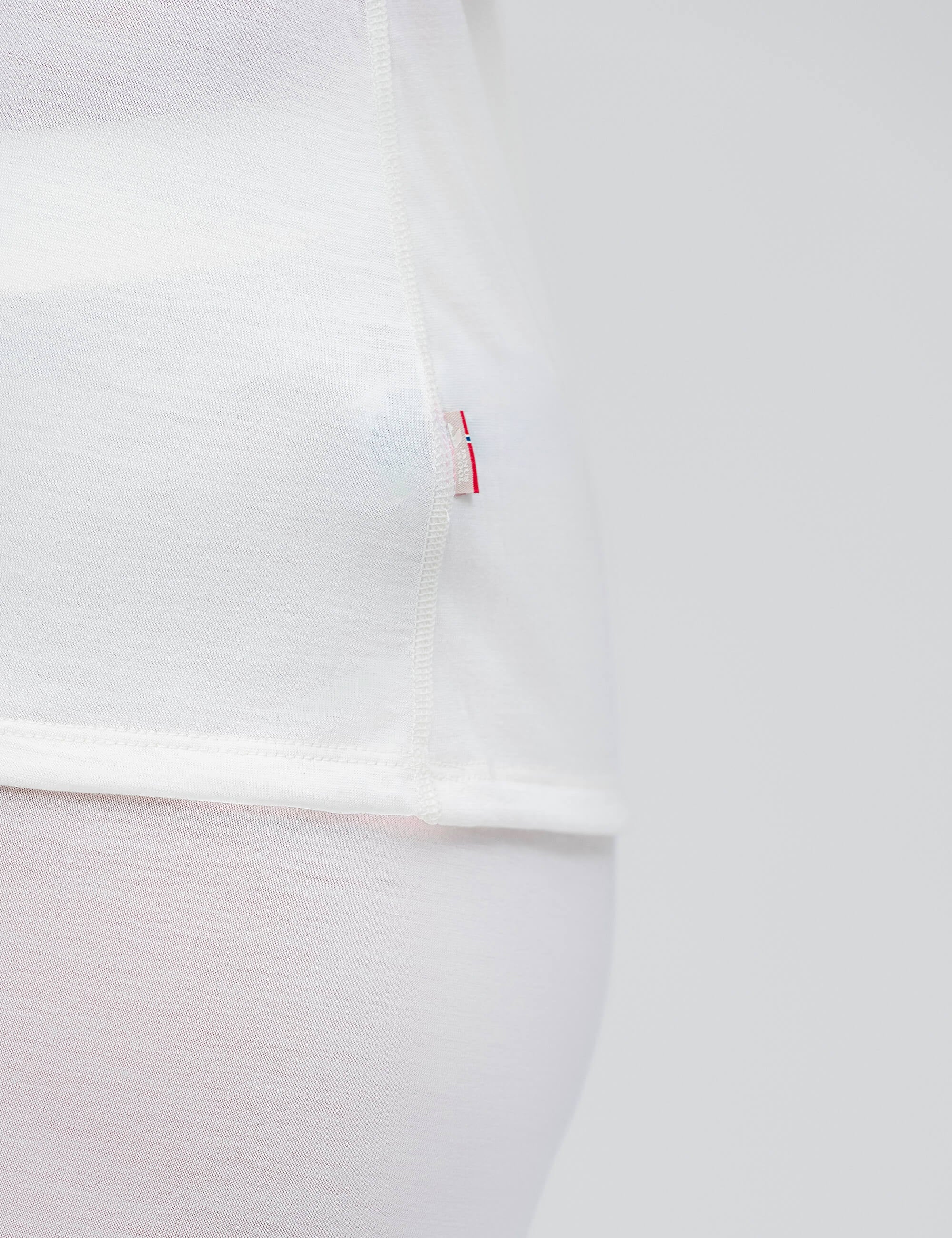 Close-up of the side seam of the Crew Neck – Classic Merino 160 Women t-shirt in white, featuring a small red and white tag. Set against a minimal all-white background for a clean, everyday style.