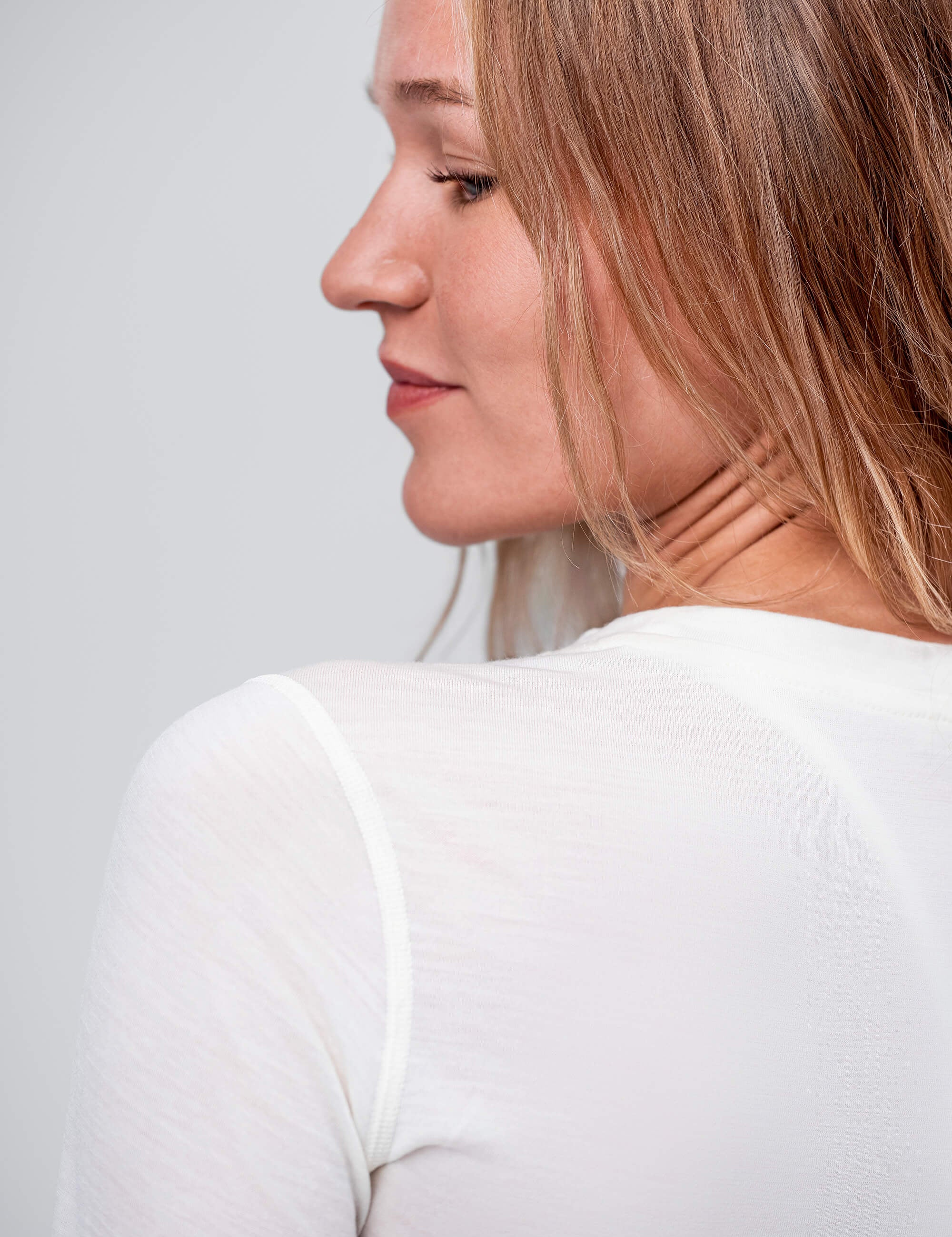 A woman with light brown hair and a peaceful expression looks over her shoulder, wearing the Crew Neck – Classic Merino 160 Women, a sheer white long-sleeve layer ideal for daily wear. The background is plain and light-colored.