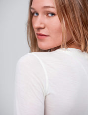 A woman with light brown hair and blue eyes looks over her shoulder, modeling the Crew Neck – Classic Merino 160 Women in sheer white, against a plain light gray background.