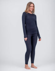 A woman stands barefoot against a plain background, wearing the Crew Neck – Classic Merino 160 Women in navy blue with matching leggings. She has light brown hair, smiles slightly, and rests her hands at her sides—ideal for everyday wear.