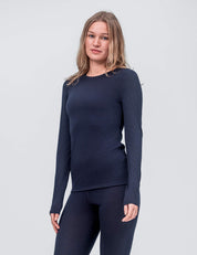 A woman with long, light brown hair wears the Crew Neck – Classic Merino 160 Women in Navy and matching leggings, an ideal everyday long-sleeve layer, while standing against a plain light grey background.
