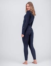 A woman with shoulder-length blonde hair, barefoot on a light gray background, smiles over her shoulder in the Crew Neck – Classic Merino 160 Women and matching leggings, highlighting this long-sleeve everyday layer.