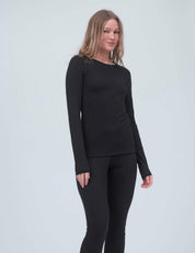 A woman with long, light brown hair is standing and smiling, wearing the Crew Neck – Classic Merino 160 Women in black and matching black leggings against a plain light gray background.