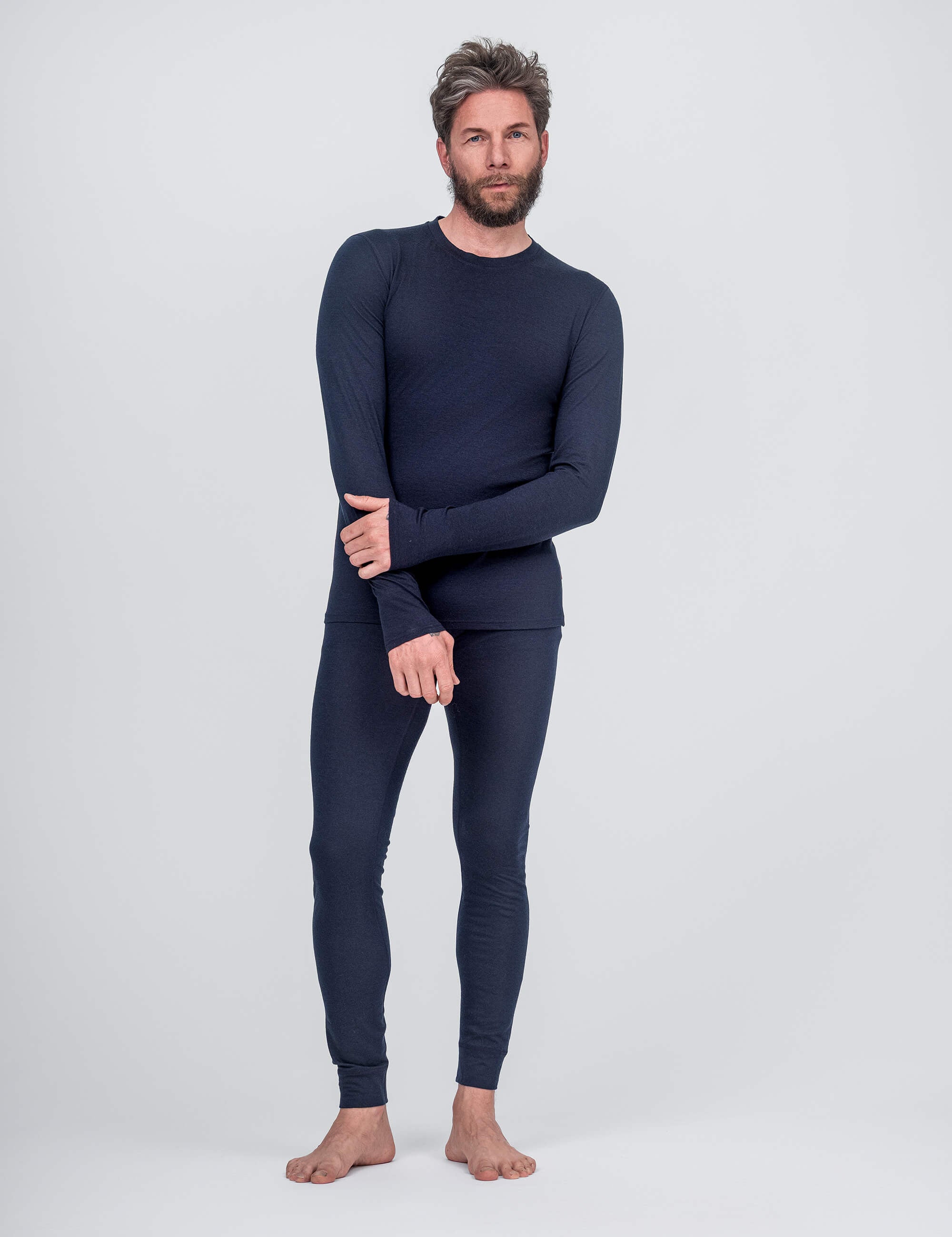 A bearded, barefoot man with messy hair stands against a plain background wearing navy Leggings – Classic Merino 160 Men and a matching thermal top, holding his opposite wrist.