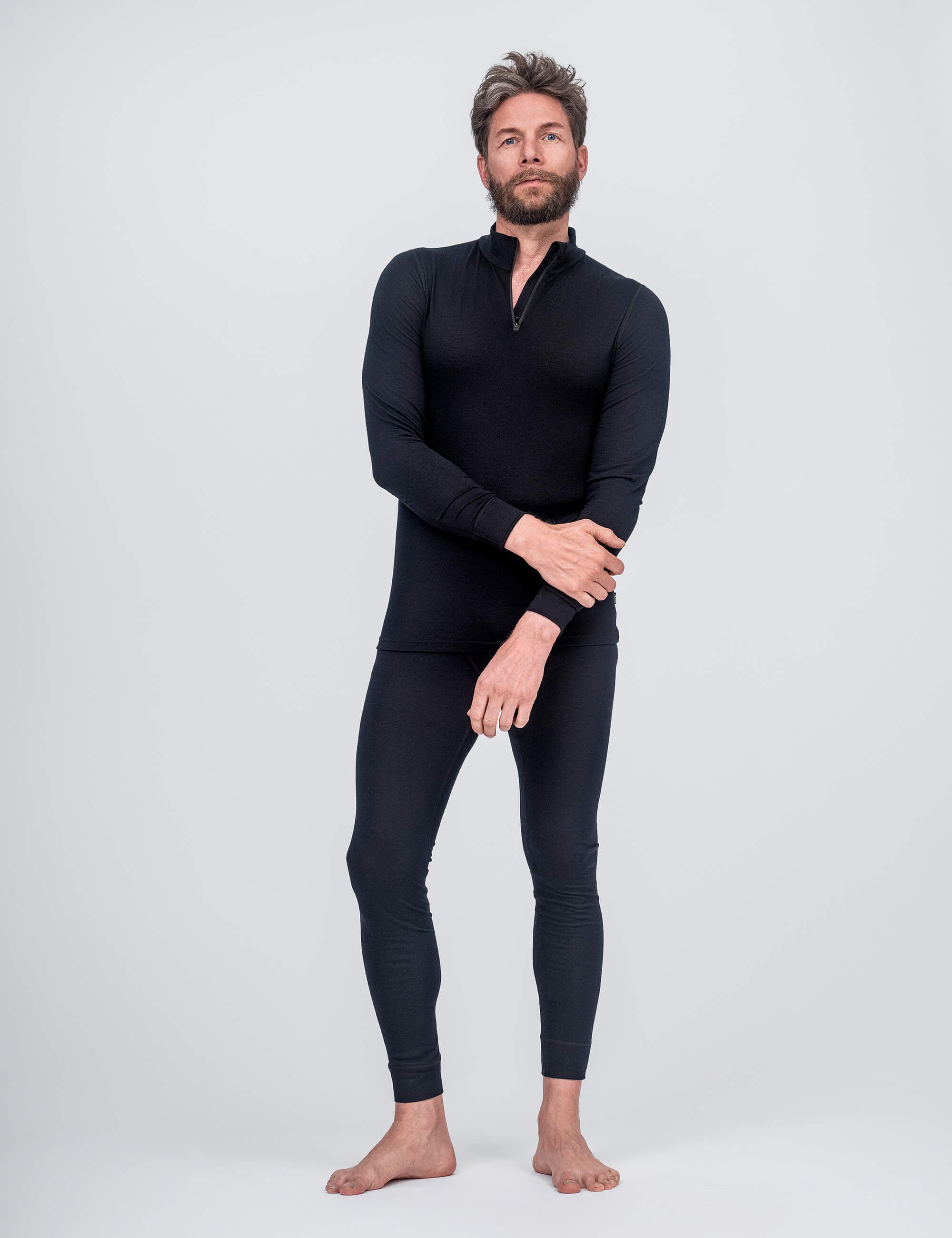 A man with short hair and a beard poses barefoot against a plain light background, wearing fitted, long-sleeve black thermal top with zip collar and Classic Merino 160 Men leggings in black.