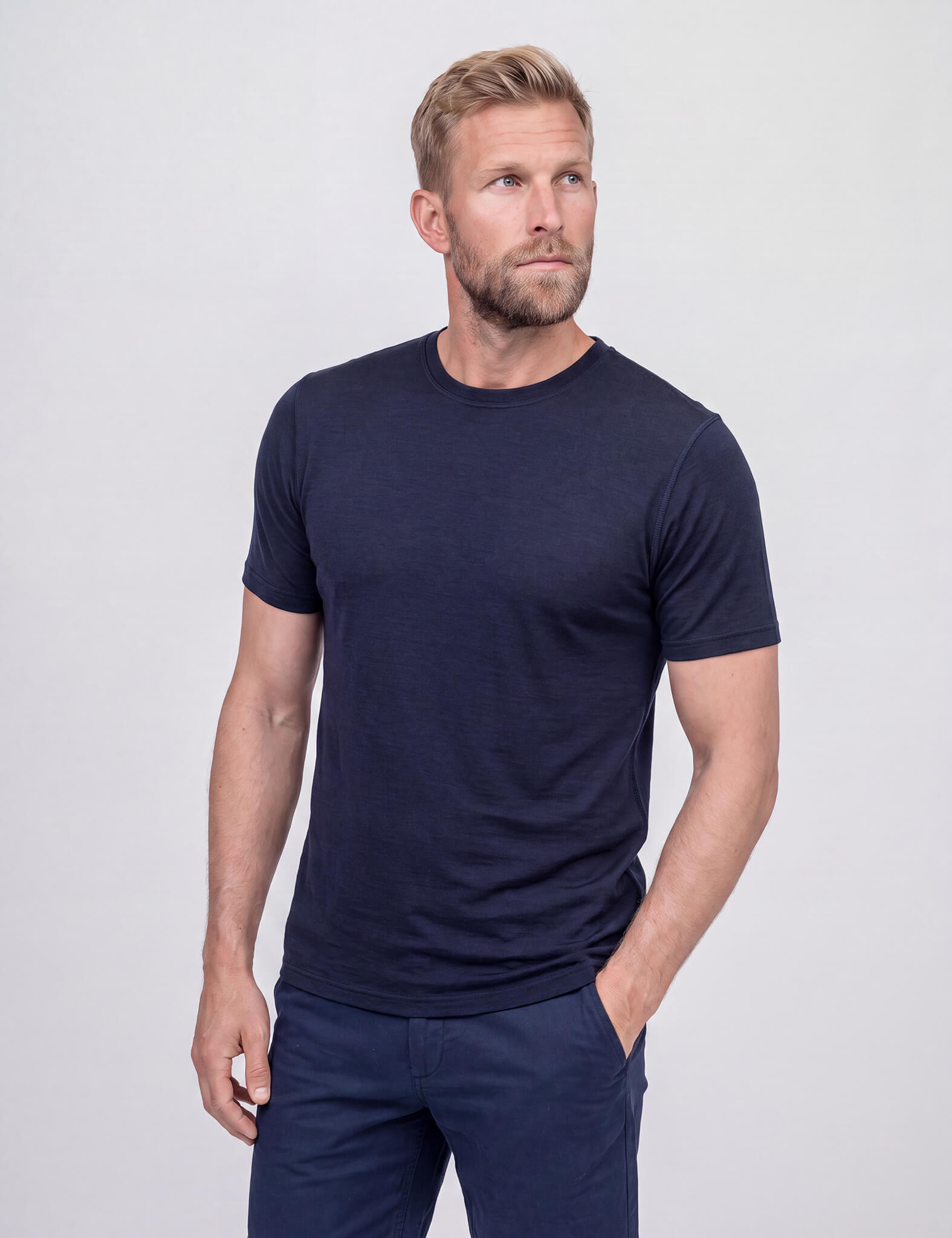 A bearded man with short hair wears the T-shirt Active – Classic Merino 160 Men, a fitted black merino shirt known for temperature regulation. He looks back and smiles slightly against a plain, light-colored background.