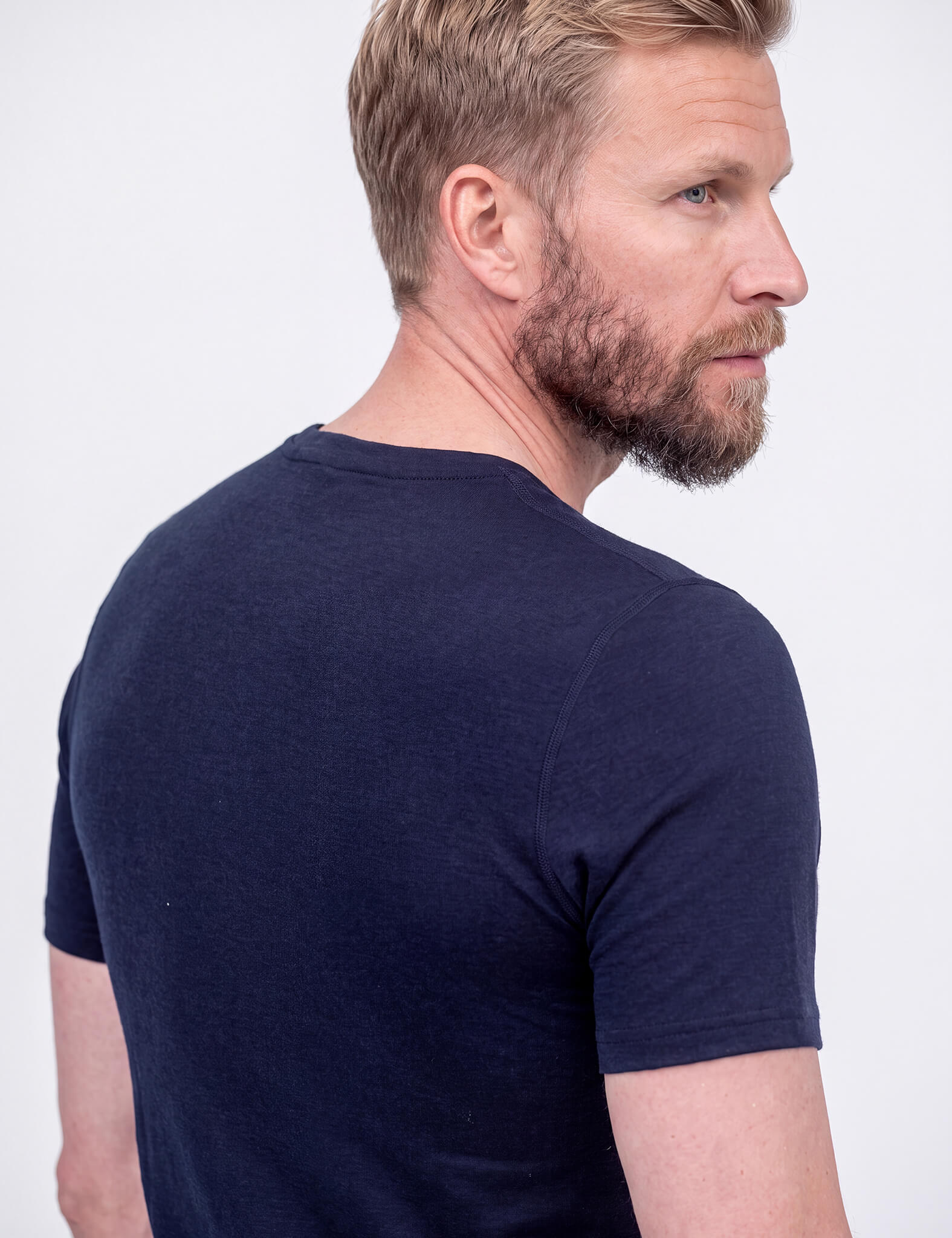 A close-up shows a bearded man in a dark blue T-shirt Active – Classic Merino 160 Men, photographed from the back and side with his head turned slightly right, set against a plain light background.