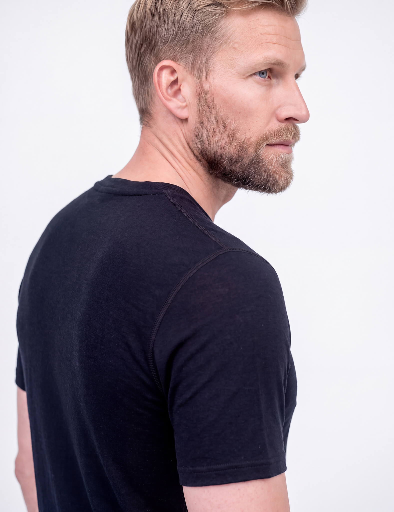 A man with light brown hair and a beard stands sideways, looking over his shoulder at the camera. He wears the T-shirt Active – Classic Merino 160 Men, paired with dark blue shorts, hand in pocket, against a plain light background.