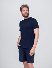 A man with a beard and short, tousled hair wears the T-shirt Regular – Classic Merino 160 Men and matching navy shorts. With one hand in his pocket, he looks to the side against a plain light gray background.
