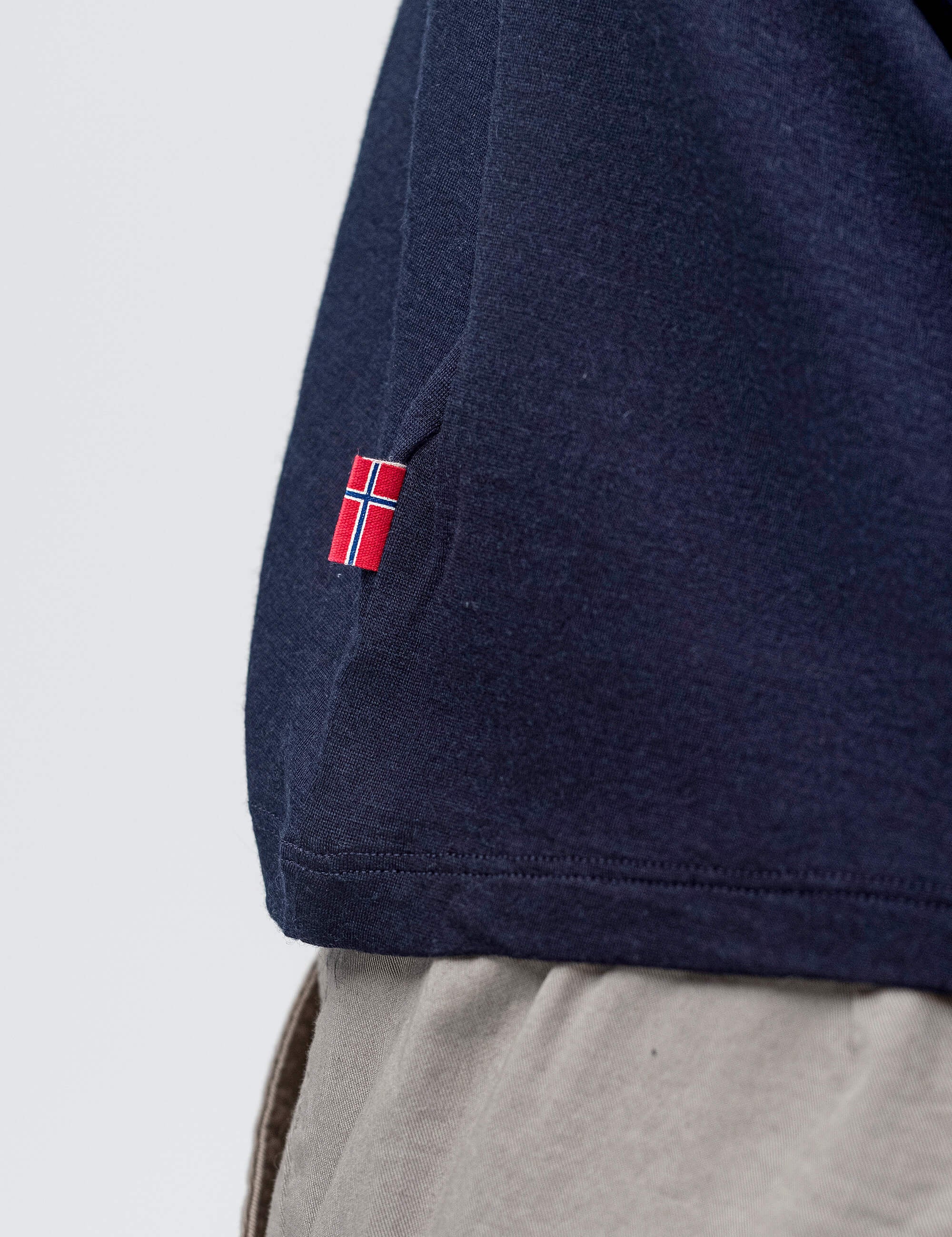 A close-up of a person wearing the T-shirt Regular – Classic Merino 160 Men in breathable navy blue merino wool with a small embroidered Norwegian flag near the hem, paired with light beige pants.