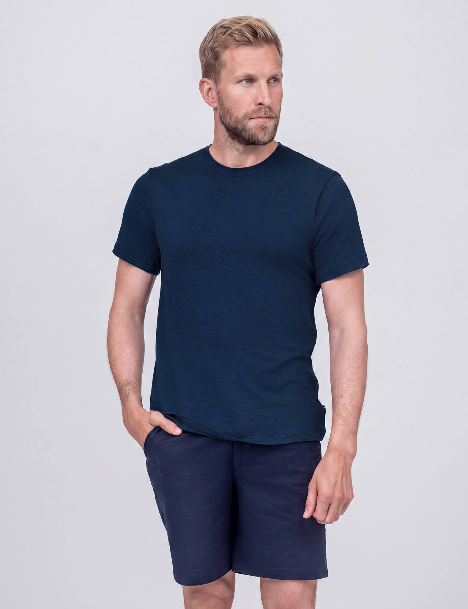 A man with wavy hair and a beard wears the Navy T-shirt Regular – Classic Merino 160 Men. He stands against a light gray background, one hand in his pocket, looking slightly to the side and smiling.