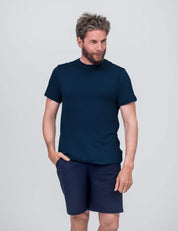 A man with wavy hair and a beard wears the Navy T-shirt Regular – Classic Merino 160 Men. He stands against a light gray background, one hand in his pocket, looking slightly to the side and smiling.