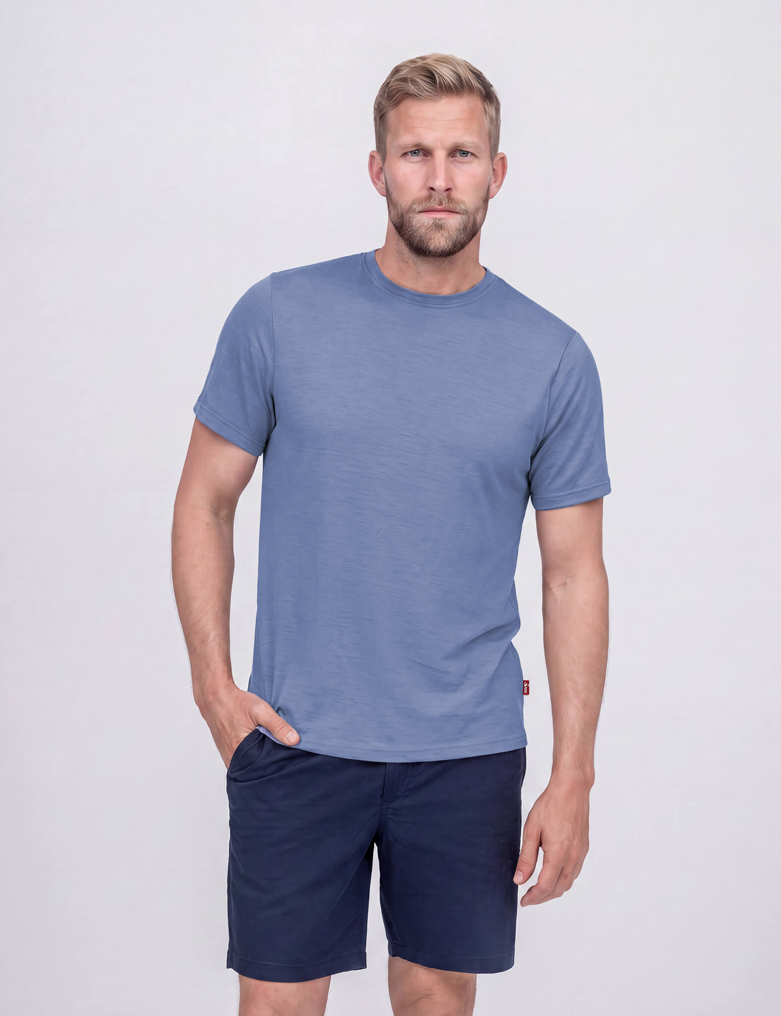 A man with short gray hair and a beard stands against a plain, light background, wearing the T-shirt Regular – Classic Merino 160 Men in Troposphere and dark blue shorts. He faces the camera with a relaxed expression.