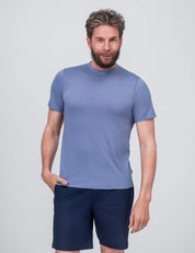 A man with short gray hair and a beard stands against a plain, light background, wearing the T-shirt Regular – Classic Merino 160 Men in Troposphere and dark blue shorts. He faces the camera with a relaxed expression.