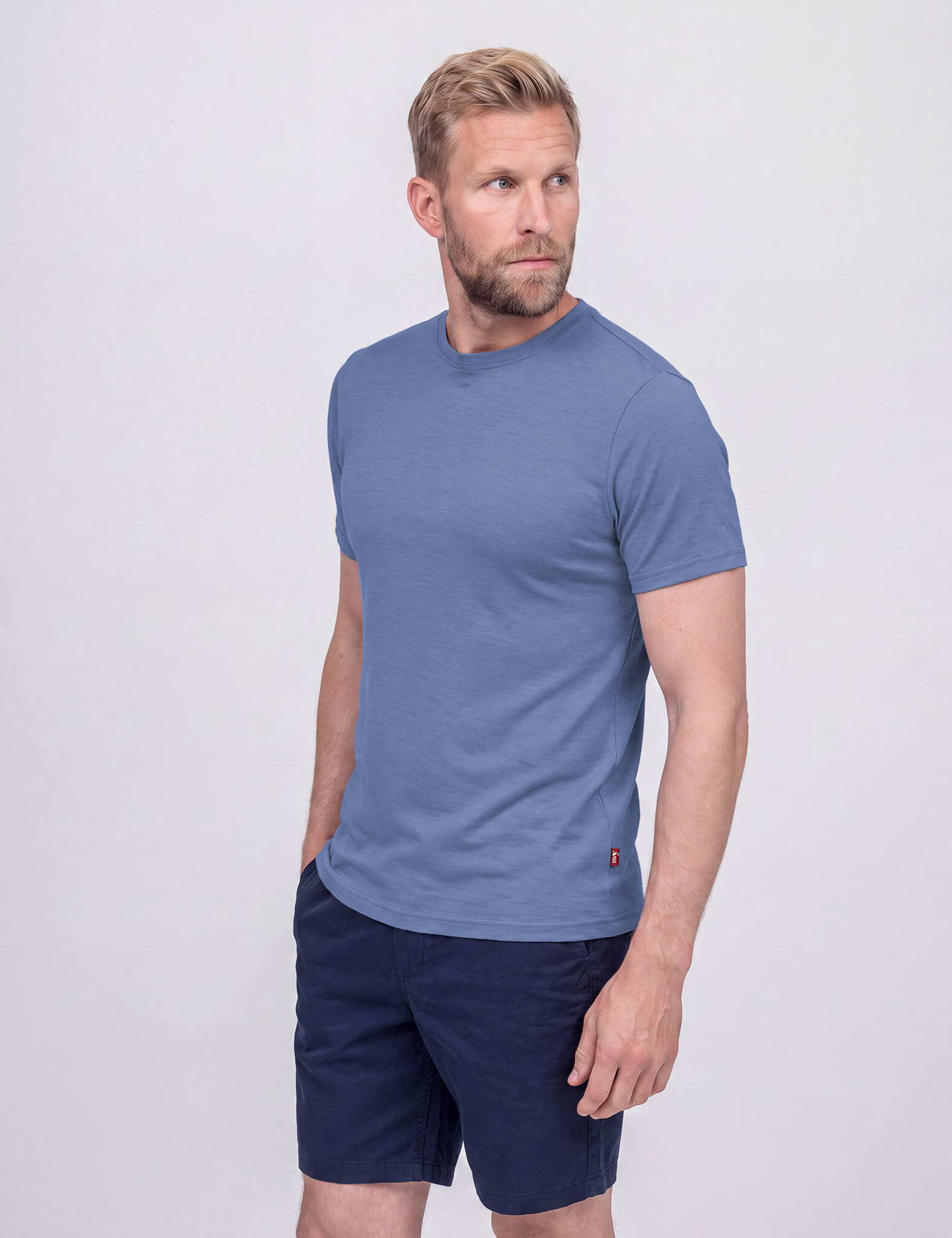 A man with short, curly hair and a beard wears the T-shirt Regular – Classic Merino 160 Men and navy shorts. He stands against a plain light background, looking left with his right hand in his pocket.