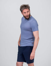 A man with short, curly hair and a beard wears the T-shirt Regular – Classic Merino 160 Men and navy shorts. He stands against a plain light background, looking left with his right hand in his pocket.