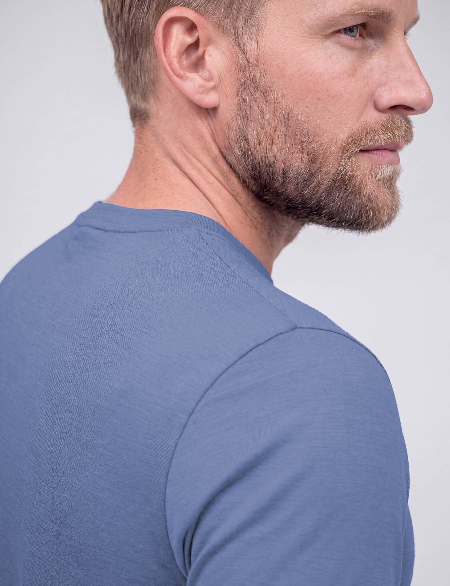 A bearded man with short hair, wearing the T-shirt Regular – Classic Merino 160 Men in light blue, stands slightly turned and subtly smiling against a light grey background.