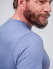 A bearded man with short hair, wearing the T-shirt Regular – Classic Merino 160 Men in light blue, stands slightly turned and subtly smiling against a light grey background.