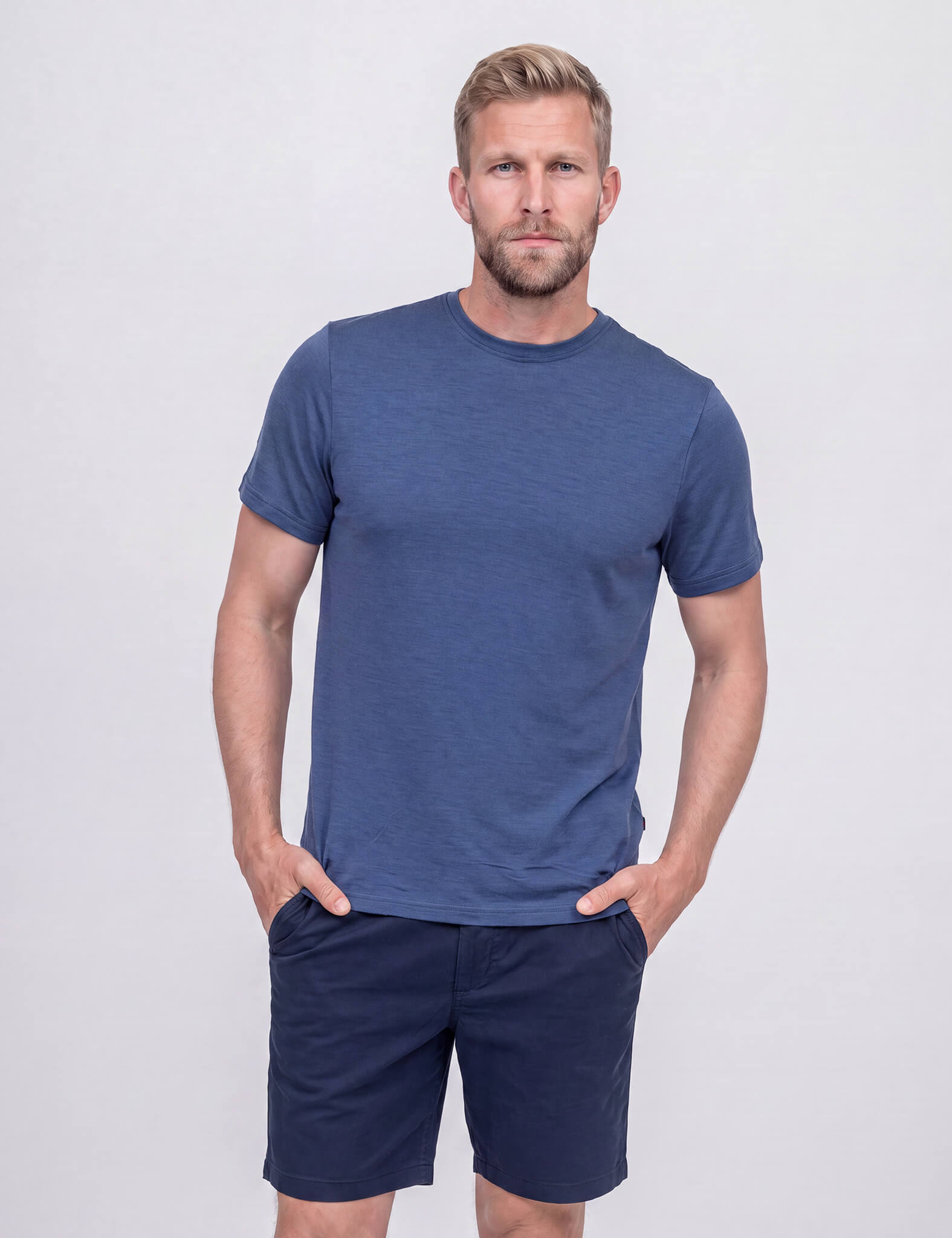 A man with short brown hair and a beard stands against a light gray background, wearing the T-shirt Regular – Classic Merino 160 Men in Ensign Blue, paired with dark blue shorts. His hands are in his pockets and he has a slight smile.