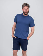 A man with short brown hair and a beard stands against a light gray background, wearing the T-shirt Regular – Classic Merino 160 Men in Ensign Blue, paired with dark blue shorts. His hands are in his pockets and he has a slight smile.