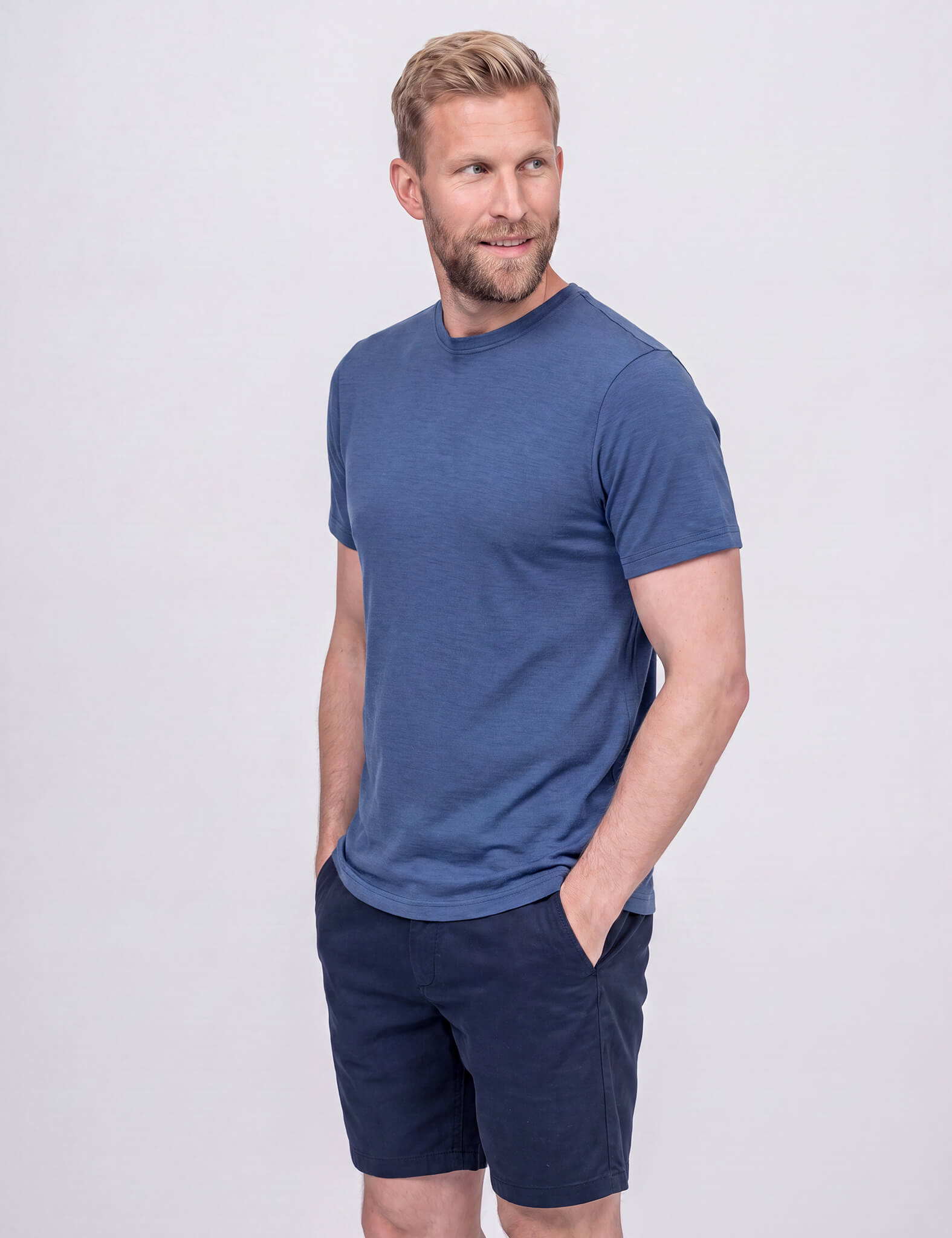 A bearded man with tousled hair wears the T-shirt Regular – Classic Merino 160 Men in blue and navy shorts, standing with hands in pockets and smiling while looking to the side against a light gray background.