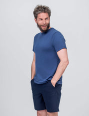 A bearded man with tousled hair wears the T-shirt Regular – Classic Merino 160 Men in blue and navy shorts, standing with hands in pockets and smiling while looking to the side against a light gray background.