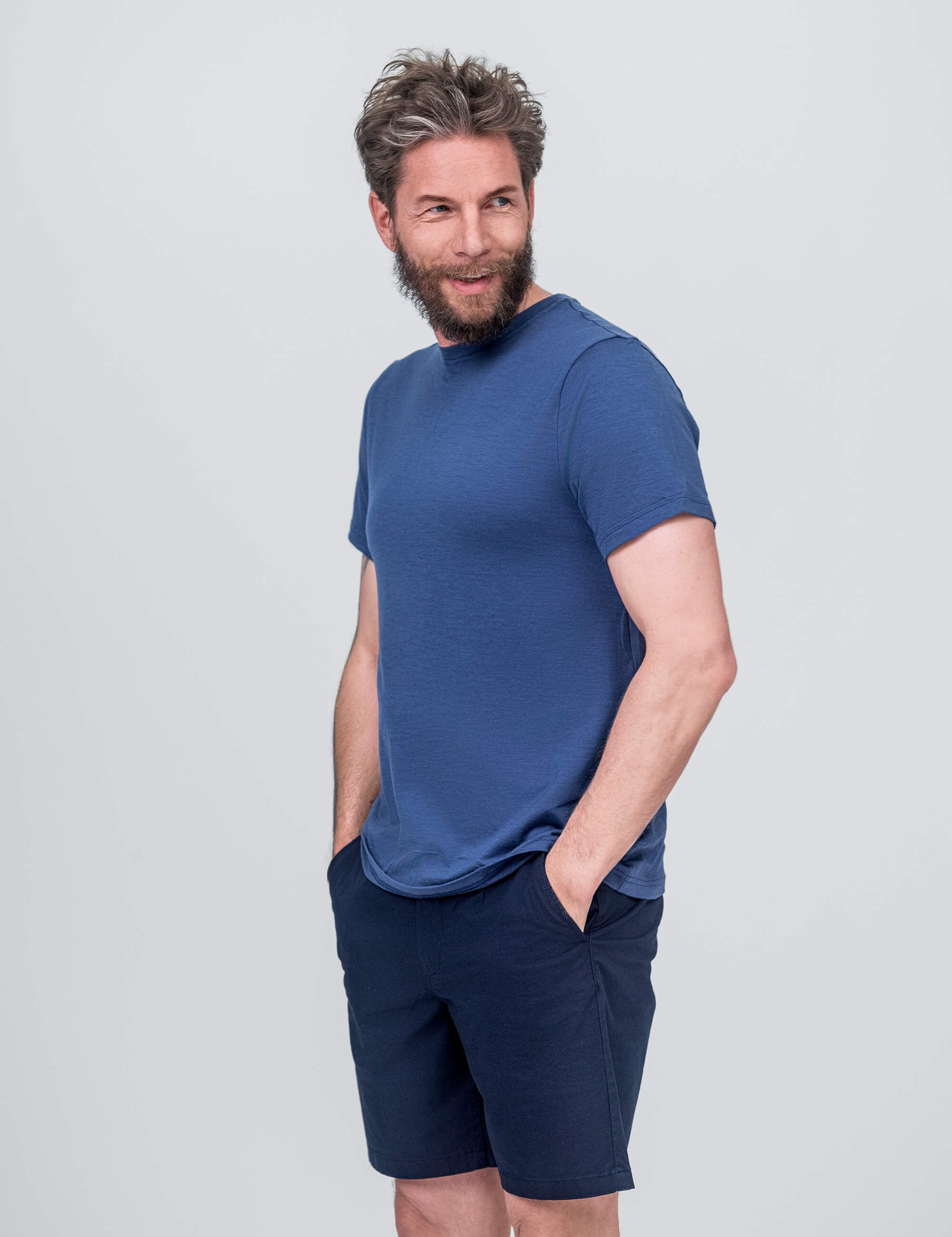 A bearded man with tousled hair wears the T-shirt Regular – Classic Merino 160 Men in blue and navy shorts, standing with hands in pockets and smiling while looking to the side against a light gray background.