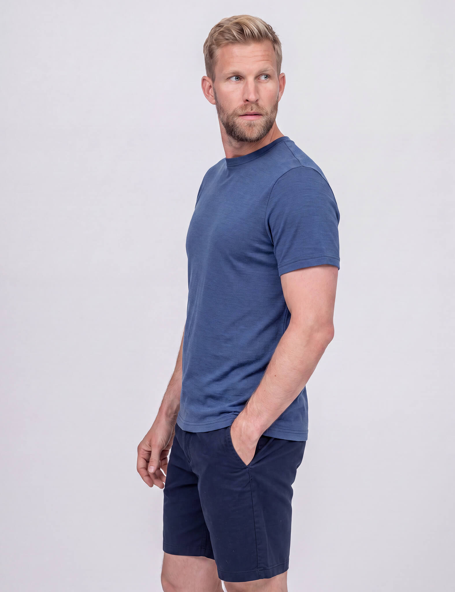 A man with light brown hair and a beard wears the T-shirt Regular – Classic Merino 160 Men and dark blue shorts. He stands sideways, one hand in his pocket, looking over his shoulder against a plain light gray background.