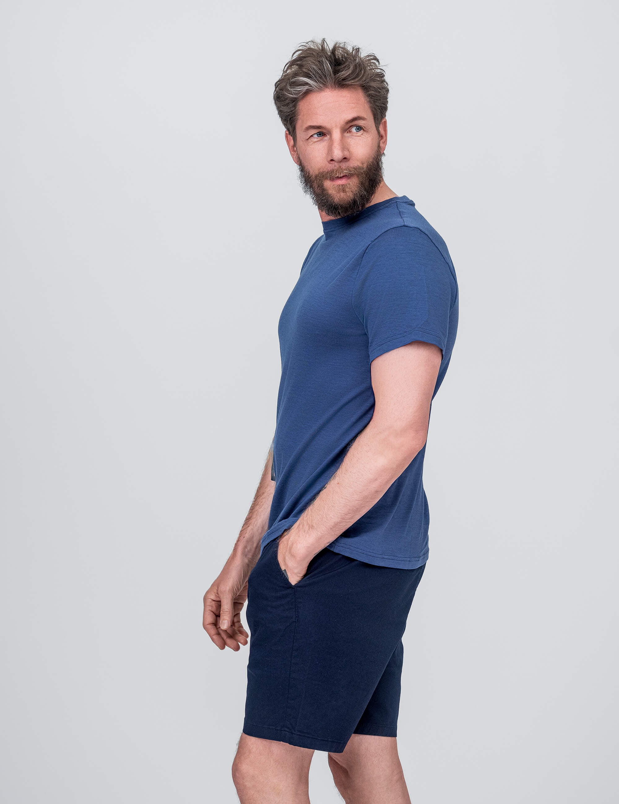 A man with light brown hair and a beard wears the T-shirt Regular – Classic Merino 160 Men and dark blue shorts. He stands sideways, one hand in his pocket, looking over his shoulder against a plain light gray background.