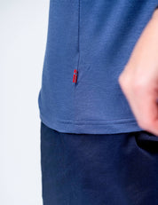 A close-up of a person wearing the T-shirt Regular – Classic Merino 160 Men in blue with dark pants. Only the torso and part of an arm are visible, and the shirt features a small red, white, and blue side seam tag. Plain background.
