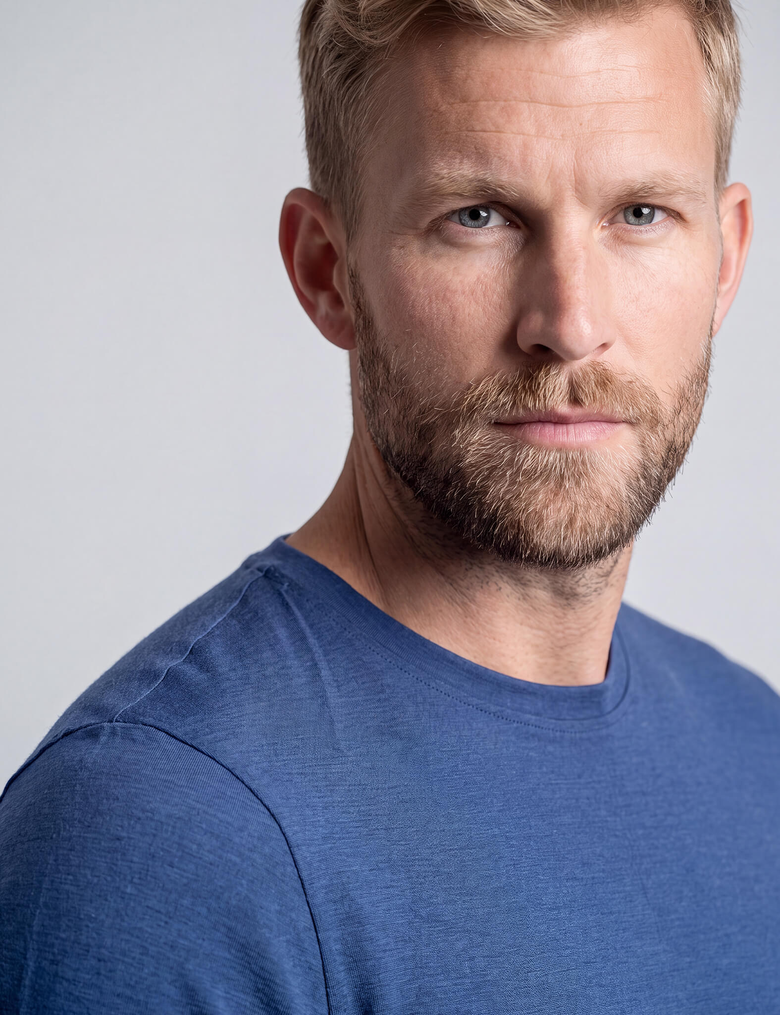 Close-up of a man with a thick beard and mustache wearing the T-shirt Regular – Classic Merino 160 Men in blue. Only the right side of his face and upper body is visible against a plain light background. He looks intently at the camera.