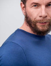 Close-up of a man with a thick beard and mustache wearing the T-shirt Regular – Classic Merino 160 Men in blue. Only the right side of his face and upper body is visible against a plain light background. He looks intently at the camera.