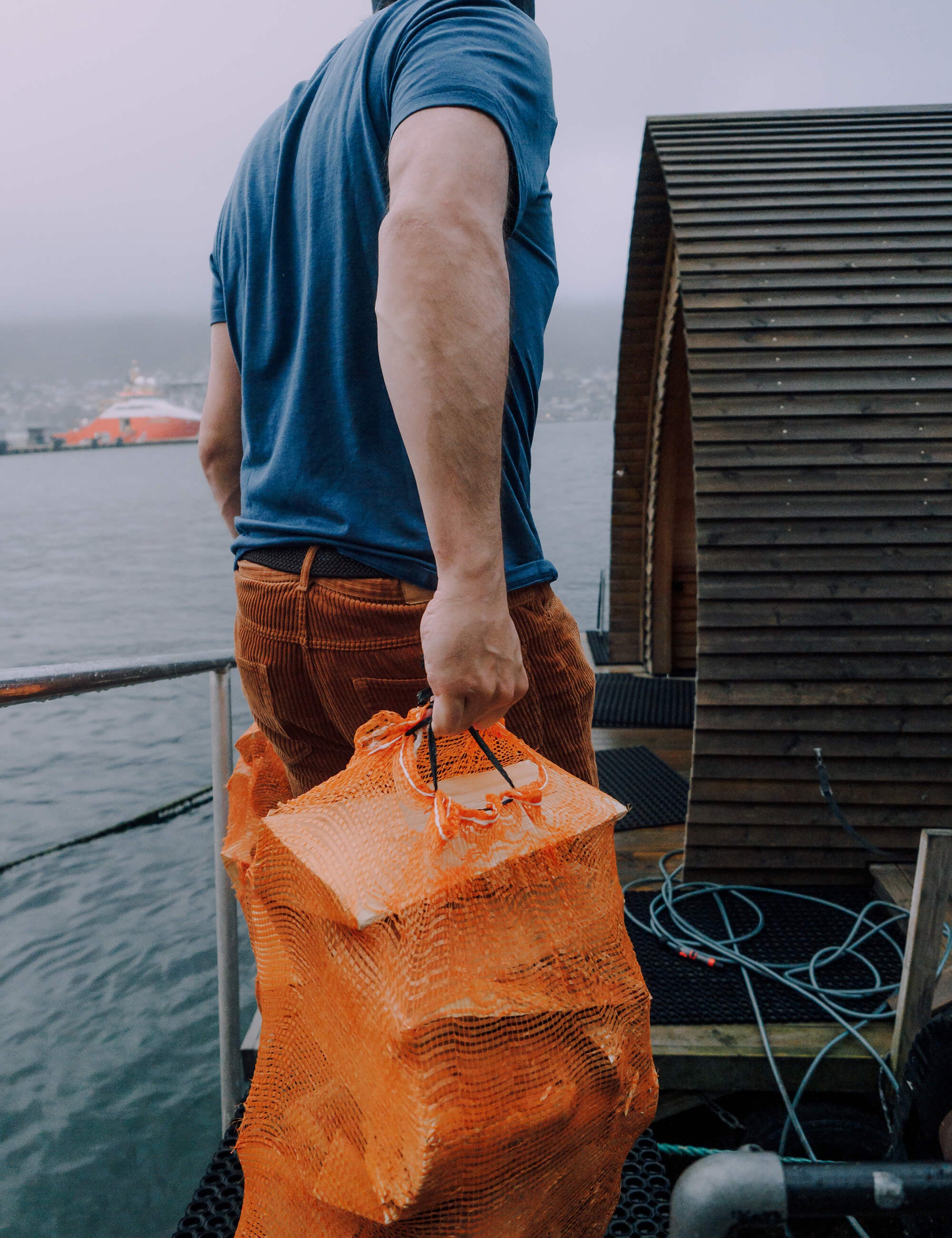 Wearing the T-shirt Regular – Classic Merino 160 Men and brown pants, a person holds an orange mesh bag while standing on a dock near a wooden structure with water and a boat in the background.
