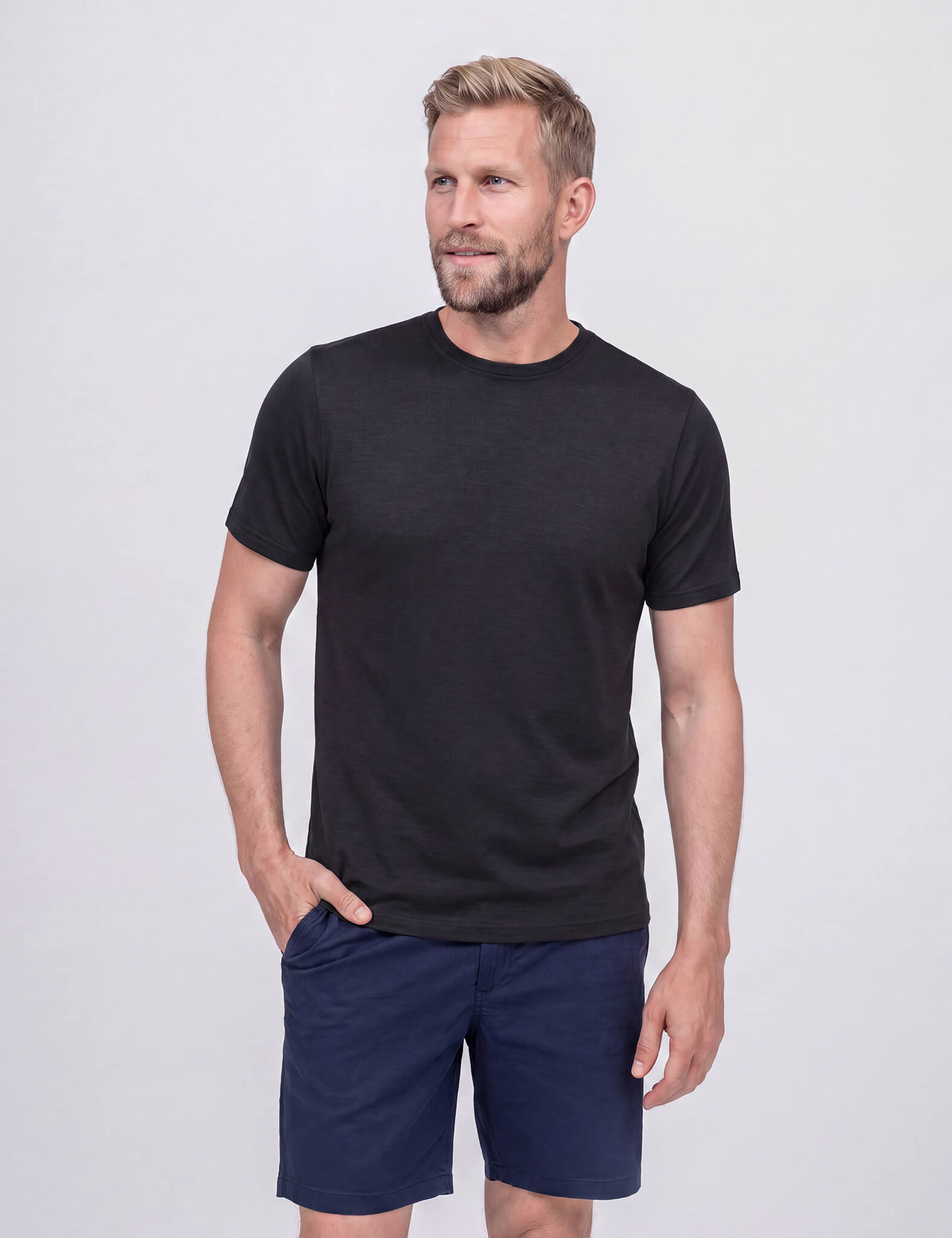 A gray-haired, bearded man smiles as he poses against a light gray background, one hand in his pocket. He wears the T-shirt Regular – Classic Merino 160 Men in black with blue shorts, looking slightly to the side.