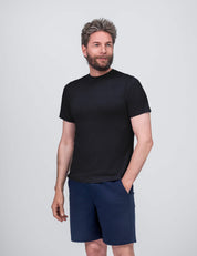 A gray-haired, bearded man smiles as he poses against a light gray background, one hand in his pocket. He wears the T-shirt Regular – Classic Merino 160 Men in black with blue shorts, looking slightly to the side.