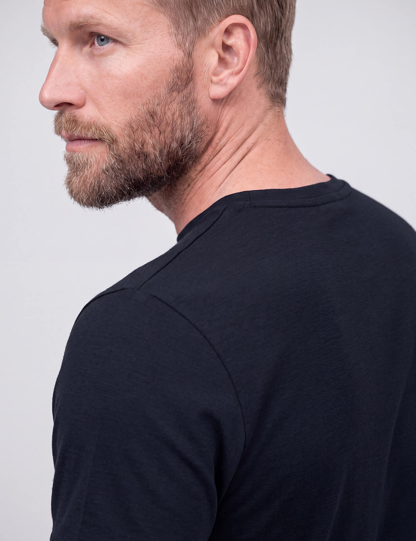 A close-up, side profile of a bearded man wearing the T-shirt Regular – Classic Merino 160 Men highlights his neck, shoulder, and part of his face against a plain light background. The breathable merino fabric offers all-day comfort.