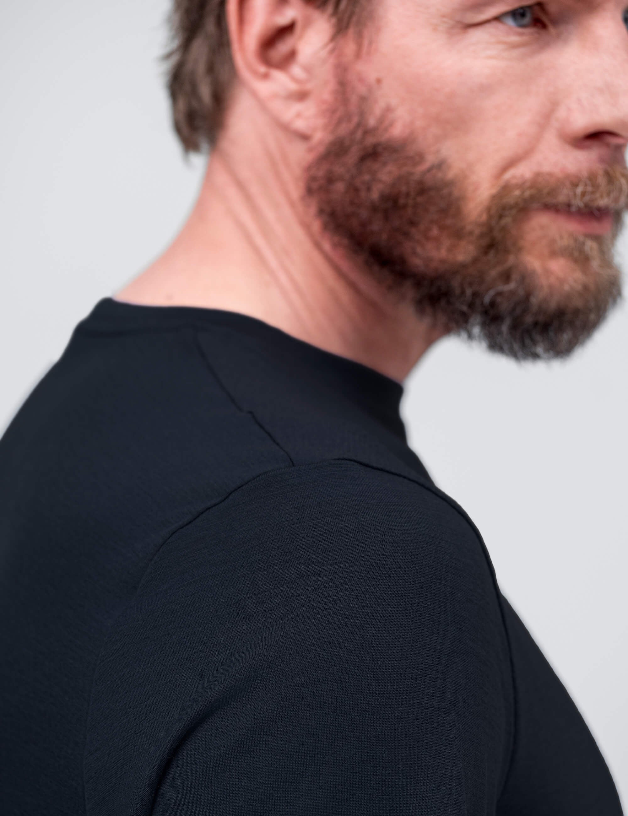 A close-up, side profile of a bearded man wearing the T-shirt Regular – Classic Merino 160 Men highlights his neck, shoulder, and part of his face against a plain light background. The breathable merino fabric offers all-day comfort.