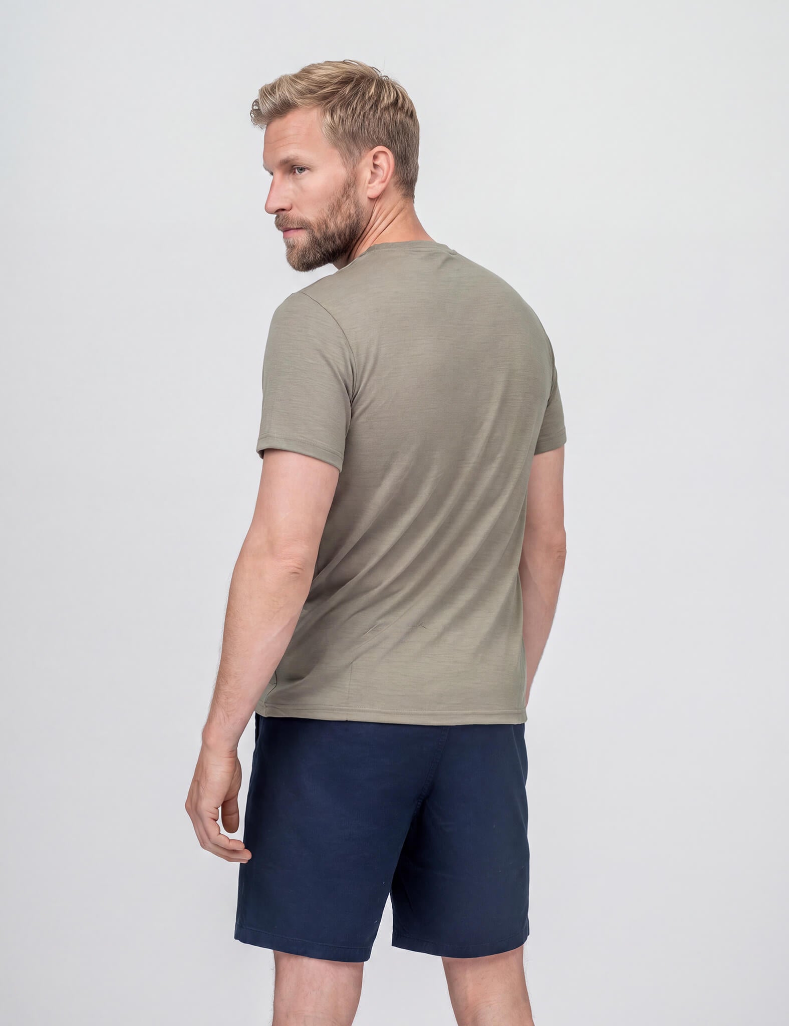 A man with short hair and a beard looks over his shoulder, wearing the T-shirt Regular – Classic Merino 160 Men in light olive green, paired with navy blue shorts against a plain light background.