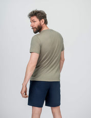 A man with short hair and a beard looks over his shoulder, wearing the T-shirt Regular – Classic Merino 160 Men in light olive green, paired with navy blue shorts against a plain light background.