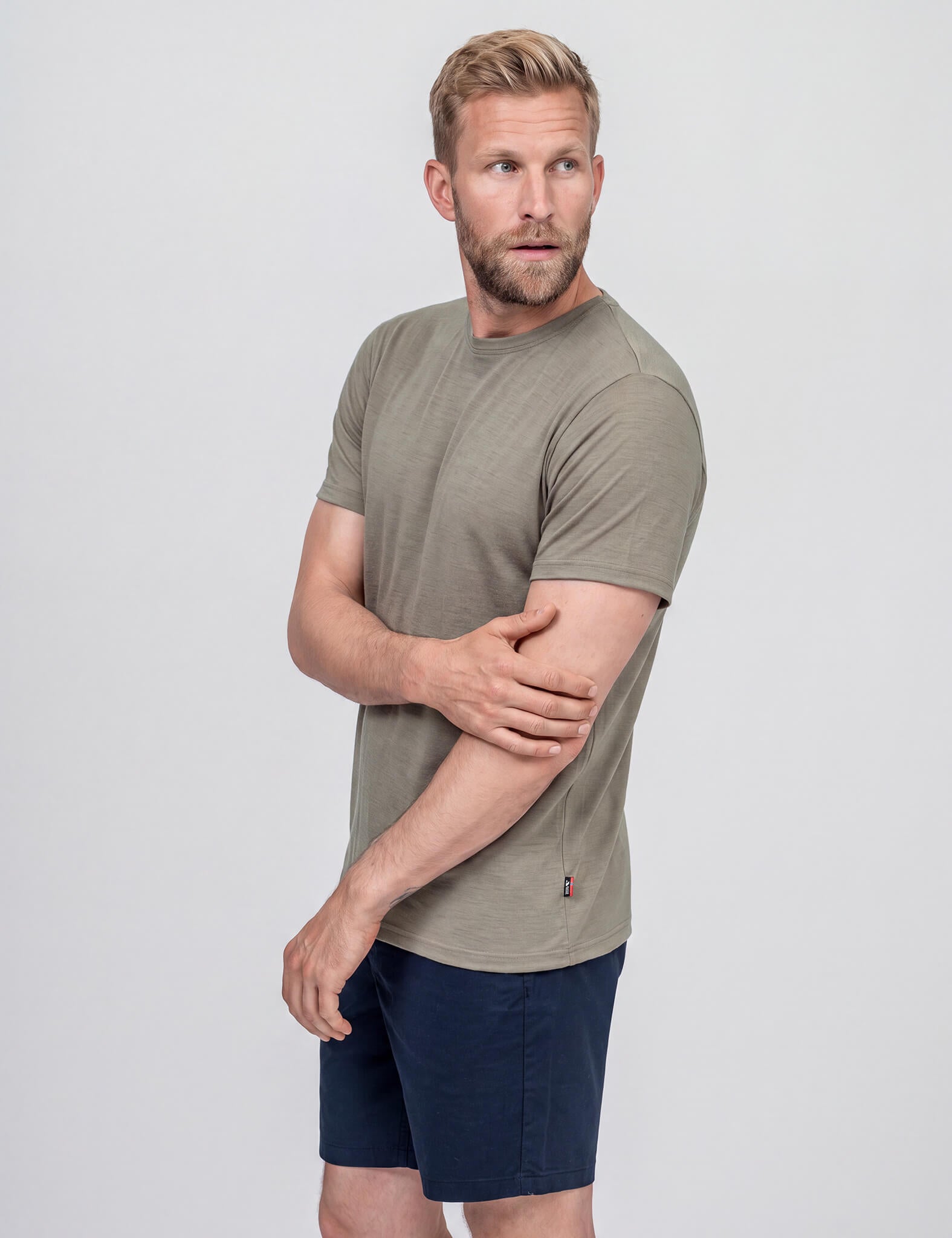 A bearded man with tousled hair wears the T-shirt Regular – Classic Merino 160 Men in Vetiver, paired with dark blue shorts, standing against a plain light gray background. He looks to his left and touches his right arm with his left hand.