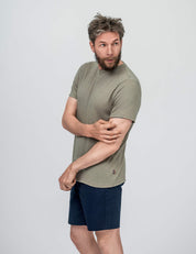 A bearded man with tousled hair wears the T-shirt Regular – Classic Merino 160 Men in Vetiver, paired with dark blue shorts, standing against a plain light gray background. He looks to his left and touches his right arm with his left hand.