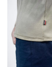 A close-up of a person wearing the T-shirt Regular – Classic Merino 160 Men in light beige, featuring a small black and red fabric tag on the side seam, paired with dark blue pants. The person's hand is partially visible in the background.