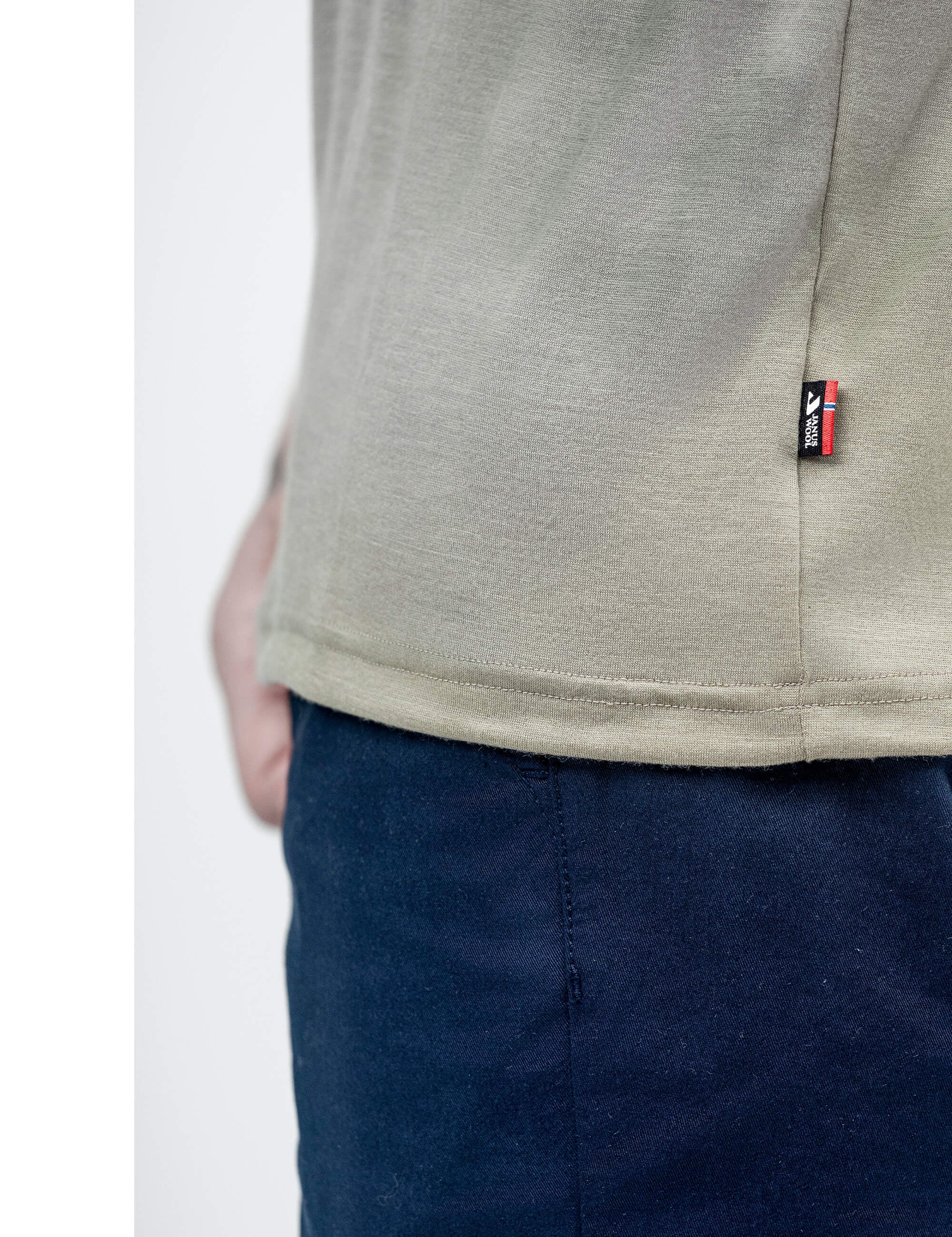 A close-up of a person wearing the T-shirt Regular – Classic Merino 160 Men in light beige, featuring a small black and red fabric tag on the side seam, paired with dark blue pants. The person's hand is partially visible in the background.