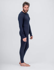 A man stands barefoot against a plain light background, wearing the Crew Neck – Classic Merino 160 Men navy thermal top and matching leggings. He faces sideways, looking toward the camera with a neutral expression.