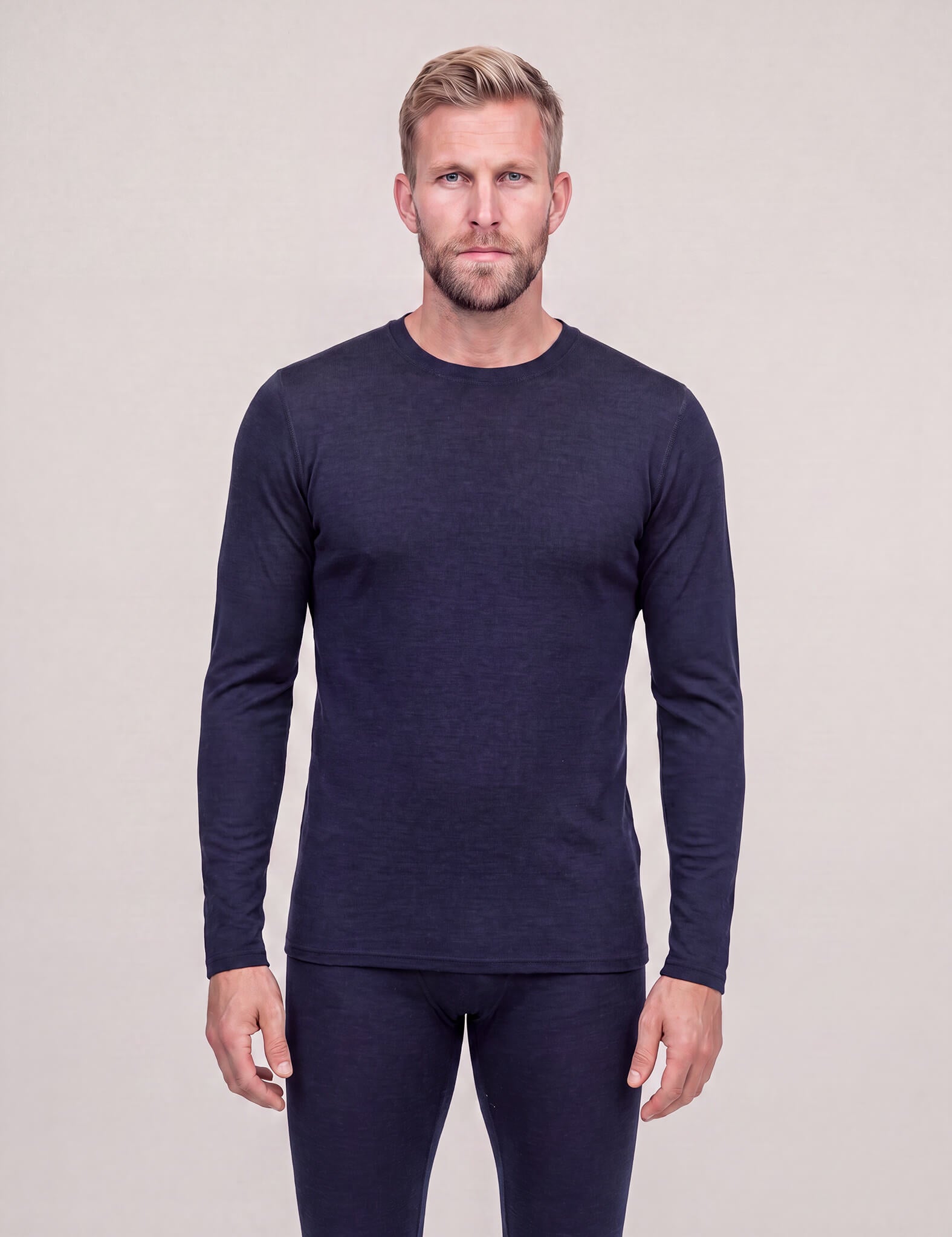 A man with blond hair and a beard is wearing the Crew Neck – Classic Merino 160 Men in navy, featuring long sleeves and matching bottoms, while standing against a plain, light background.