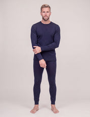 A man stands barefoot against a plain background, wearing the Crew Neck – Classic Merino 160 Men long-sleeve layer and matching fitted pants, with one hand resting on his opposite arm.