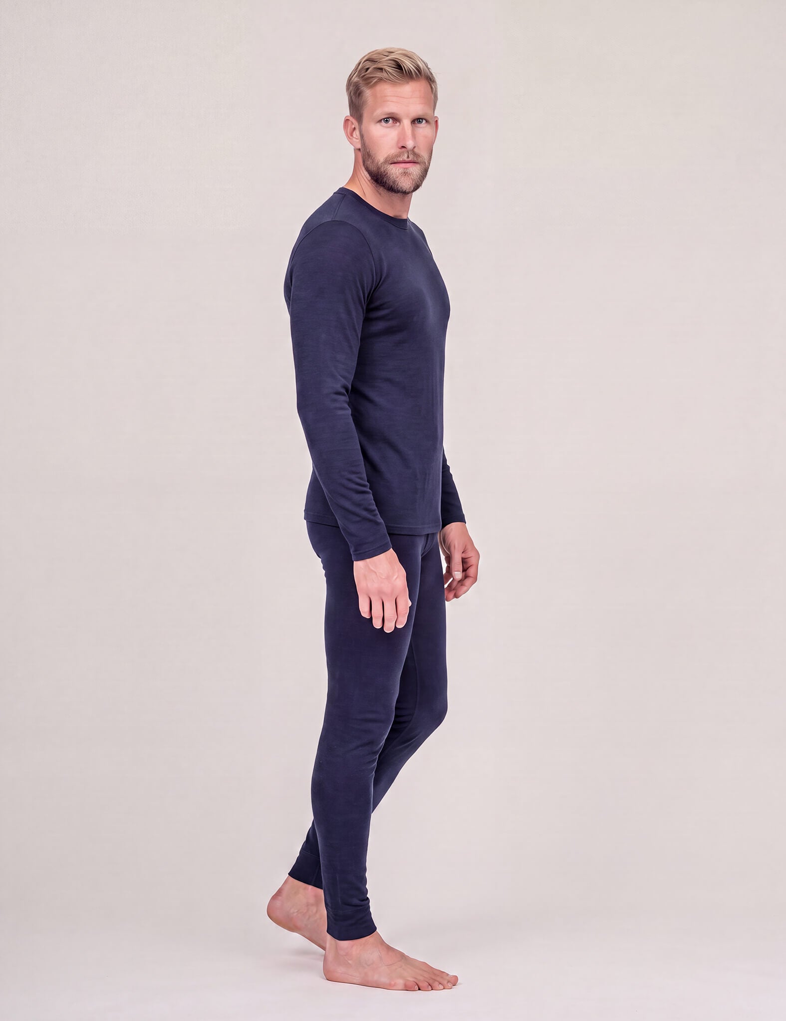 A man with short blond hair stands barefoot in profile against a light gray background, wearing the Crew Neck – Classic Merino 160 Men top in navy with matching leggings—an ideal travel-friendly layer.