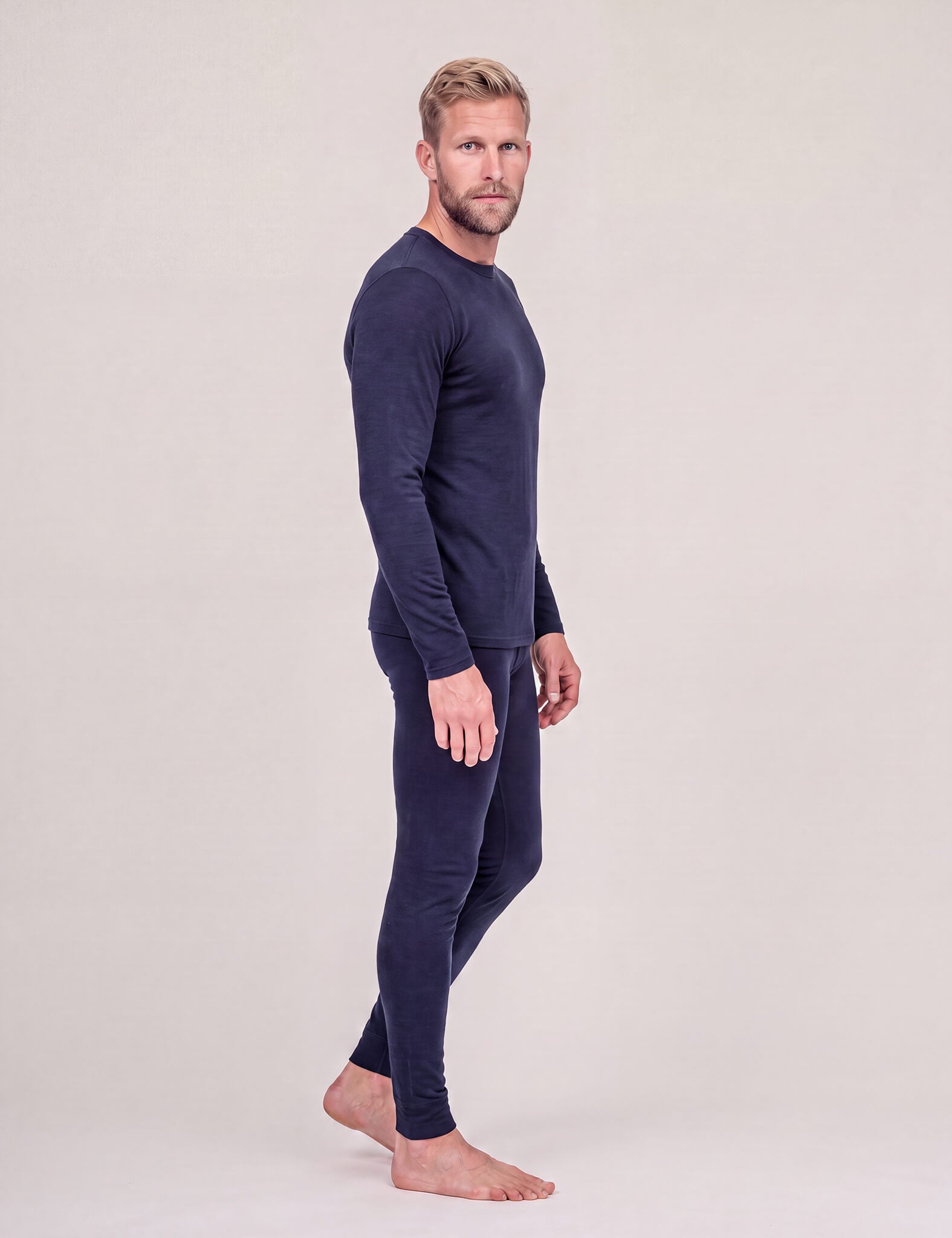 A man with short blond hair stands barefoot in profile against a light gray background, wearing the Crew Neck – Classic Merino 160 Men top in navy with matching leggings—an ideal travel-friendly layer.