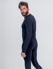 A man with a beard and tousled hair models the Classic Merino 160 Men crew neck in fitted navy blue with matching pants, standing sideways and glancing back against a plain light backdrop. Ideal for everyday wear.